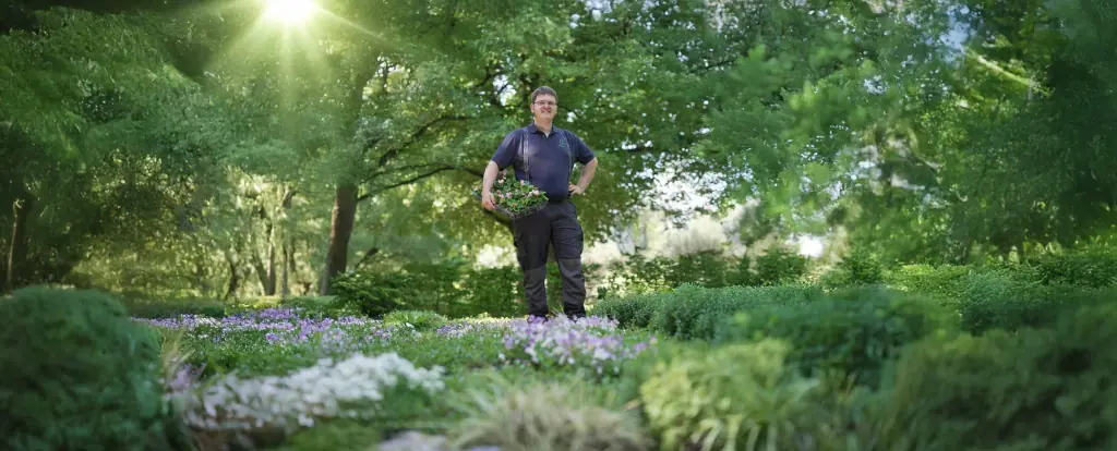 Johannes Siegele steht mit einer Pflanzschale in einem blühenden Garten unter hohen Bäumen